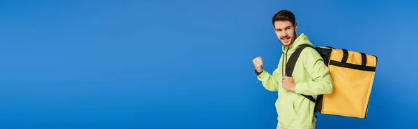 Tiro panorámico del hombre de entrega feliz mostrando gesto ganador mientras lleva la mochila térmica aislada en azul - foto de stock