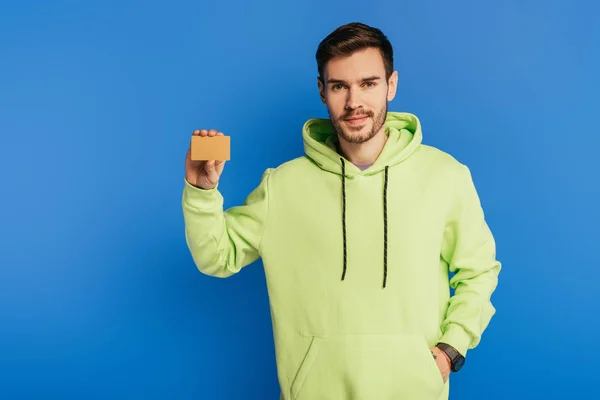 Sonriente joven mostrando tarjeta de crédito mientras sostiene la mano en el bolsillo sobre fondo azul - foto de stock