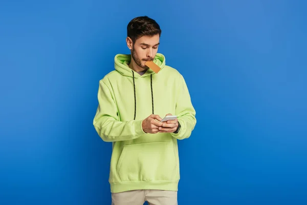 Concentrated young man using smartphone while holding credit card isolated on blue — Stock Photo