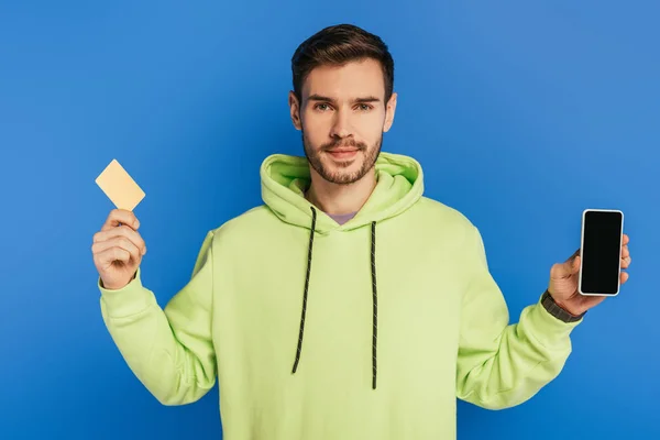 Positive young man showing smartphone with blank screen and credit card isolated on blue — Stock Photo