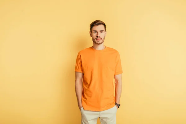 Confident young man looking at camera while holding hands in pockets on yellow background — Stock Photo