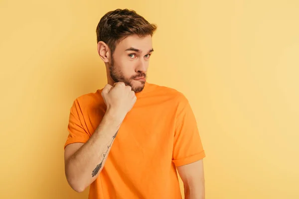 Angry young man threatening while holding fist near jaw on yellow background — Stock Photo