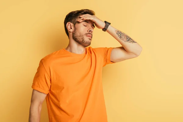 Agotado joven tocando la frente mientras está de pie con los ojos cerrados sobre el fondo amarillo - foto de stock