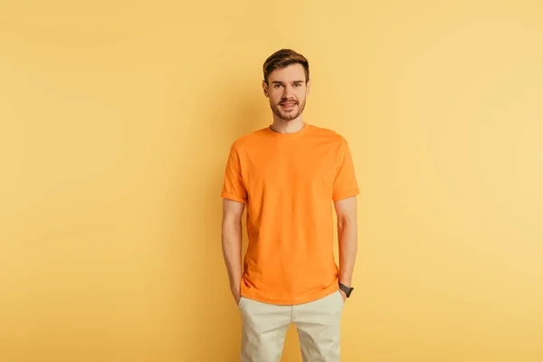 Jovem sorridente em camiseta laranja de pé com as mãos nos bolsos no fundo amarelo — Fotografia de Stock