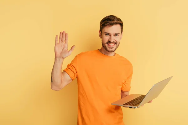 Homem feliz acenando mão na câmera enquanto segurando laptop no fundo amarelo — Fotografia de Stock