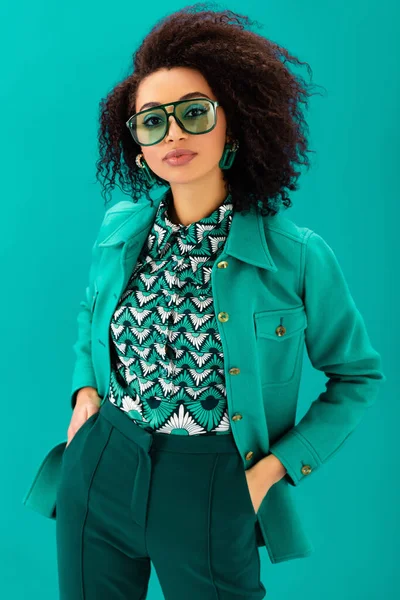 African american woman in jacket with hands in pockets looking at camera isolated on turquoise — Stock Photo