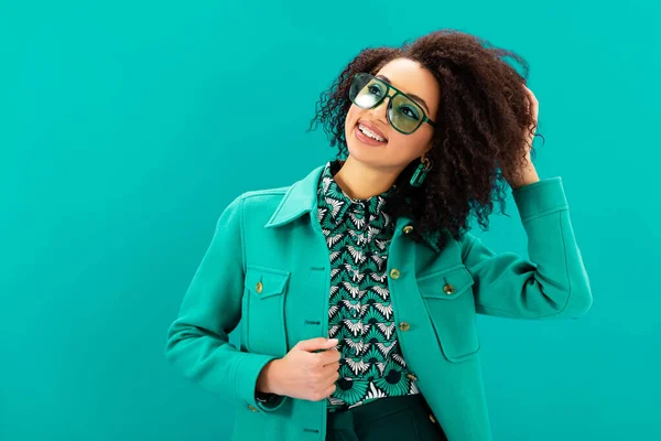 Sorridente mulher afro-americana em casaco olhando para longe isolado em turquesa — Fotografia de Stock