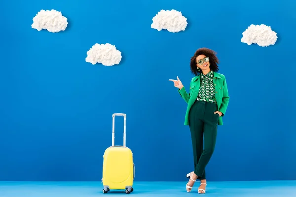 Lächelnde Afroamerikanerin zeigt mit dem Finger und steht neben Reisetasche auf blauem Hintergrund mit Wolken — Stockfoto