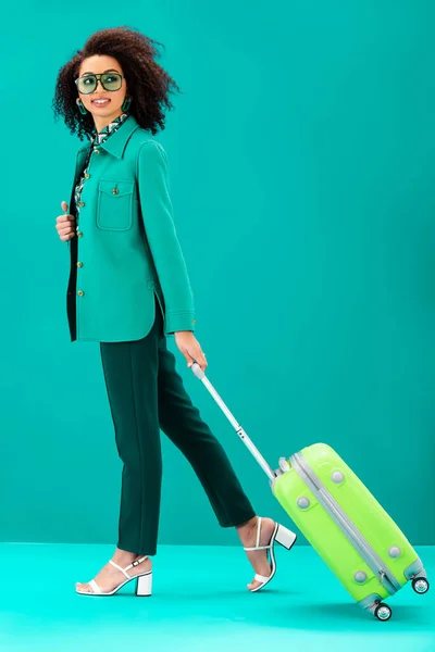 Smiling african american woman holding travel bag on turquoise background — Stock Photo