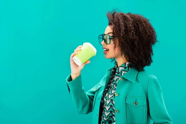 Vista lateral de la elegante mujer afroamericana bebiendo café aislado en turquesa - foto de stock