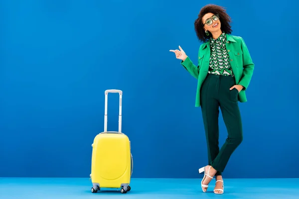 Sonriente mujer afroamericana señalando con el dedo y de pie cerca de la bolsa de viaje sobre fondo azul - foto de stock