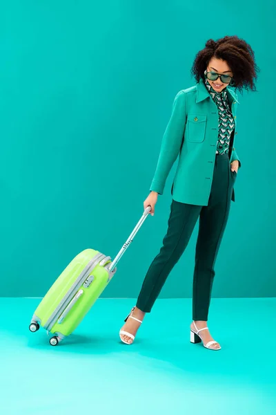 Smiling african american woman walking with travel bag on turquoise background — Stock Photo