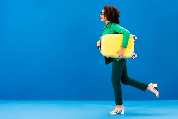 Seitenansicht einer lächelnden Afroamerikanerin, die mit Reisetasche auf blauem Grund läuft — Stockfoto