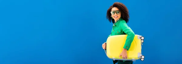 Plano panorámico de la sonriente mujer afroamericana sosteniendo bolsa de viaje aislada en azul - foto de stock