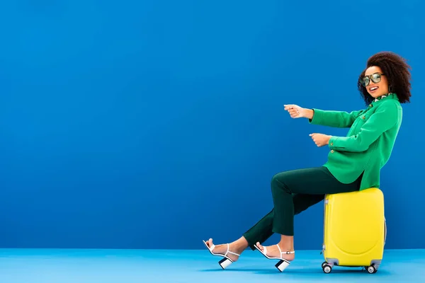 Sonriente mujer afroamericana tirando y sentado en bolsa de viaje sobre fondo azul — Stock Photo