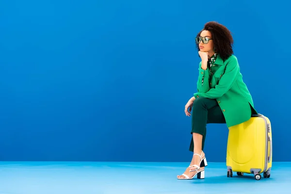 Pensive african american woman sitting on travel bag on blue background — Stock Photo