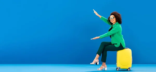 Panoramic shot of smiling african american woman sitting on travel bag and pointing with hands on blue background — Stock Photo