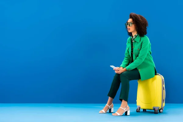 Shocked african american woman sitting on travel bag and holding smartphone on blue background — Stock Photo