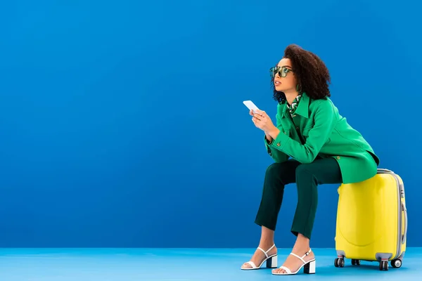 Mujer afroamericana sentada en una bolsa de viaje y sosteniendo un teléfono inteligente sobre fondo azul - foto de stock