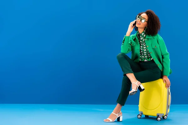 Mujer afroamericana sonriente sentada en una bolsa de viaje y hablando en un teléfono inteligente sobre fondo azul - foto de stock