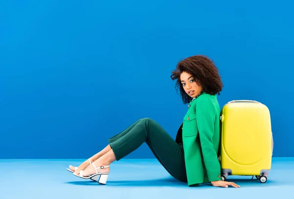 Stylish african american woman sitting near travel bag on blue background — Stock Photo