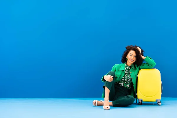 Donna afro-americana sorridente seduta vicino alla borsa da viaggio su sfondo blu — Foto stock