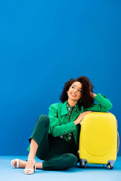 Femme afro-américaine souriante assise près d'un sac de voyage sur fond bleu — Photo de stock