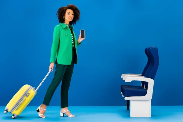 Femme afro-américaine souriante tenant sac de voyage et smartphone sur fond bleu — Photo de stock