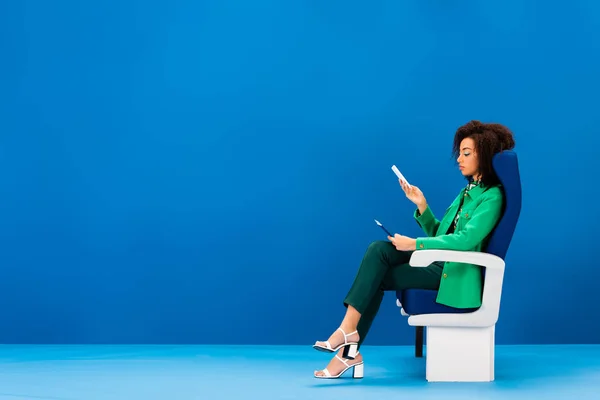 Side view of african american woman sitting on seat and holding smartphone on blue background — Stock Photo