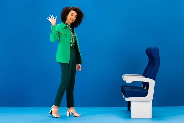 Sorridente mulher afro-americana acenando e em pé perto de assentos no fundo azul — Fotografia de Stock