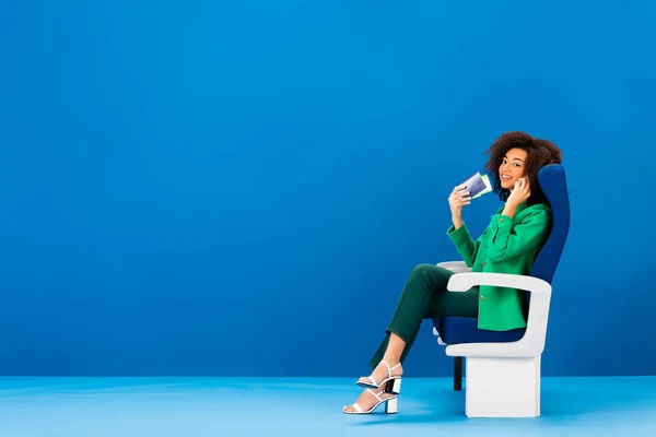 Smiling african american woman talking on smartphone and sitting on seat on blue background — Stock Photo