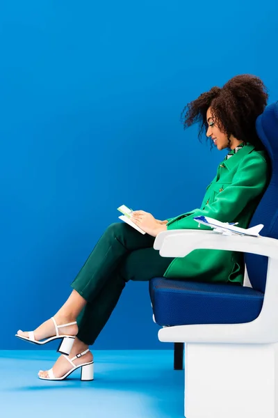 Vista lateral da mulher americana africana sorridente e modelo de avião no assento no fundo azul — Fotografia de Stock