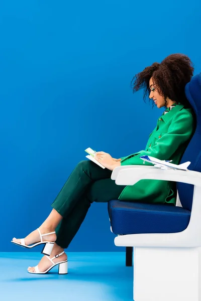 Vue latérale de la femme afro-américaine souriante et modèle d'avion sur siège sur fond bleu — Photo de stock