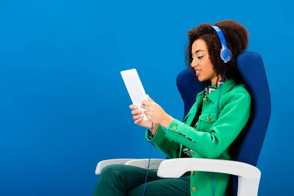 Vue latérale de sourire afro-américain en utilisant une tablette numérique et en écoutant de la musique isolée sur bleu — Photo de stock