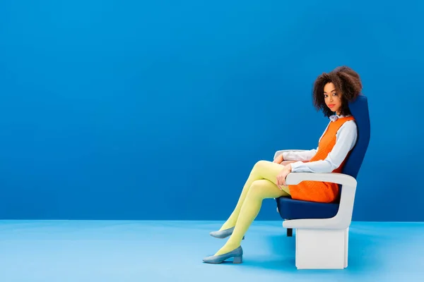 Afro-américaine en robe rétro assise sur un siège sur fond bleu — Photo de stock