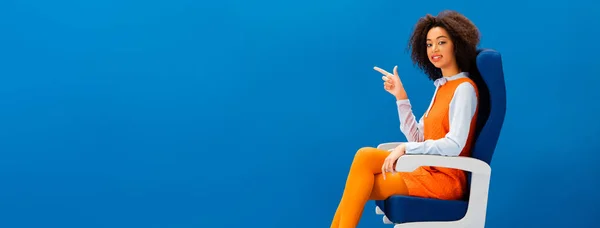 Panoramic shot of smiling african american in retro dress sitting on seat and pointing with finger isolated on blue — Stock Photo