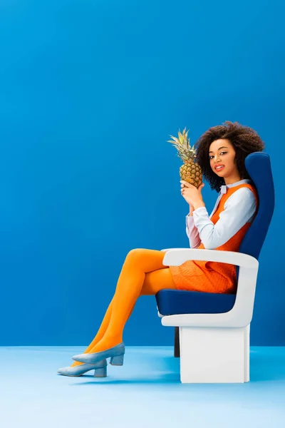 Smiling african american in retro dress sitting on seat and holding pineapple on blue background — Stock Photo