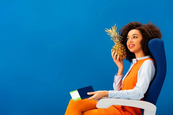 Souriant afro-américain en robe rétro tenant l'ananas et passeport avec billet d'avion isolé sur bleu — Photo de stock