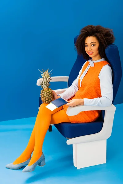 Souriant afro-américain en robe rétro tenant l'ananas et tenant passeport avec billet d'avion sur fond bleu — Photo de stock