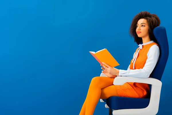 Smiling african american in retro dress sitting on seat and holding book isolated on blue — Stock Photo