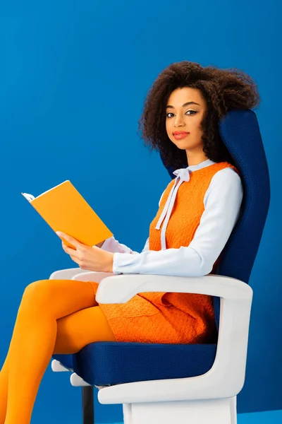 Smiling african american in retro dress holding book and sitting on seat isolated on blue — Stock Photo
