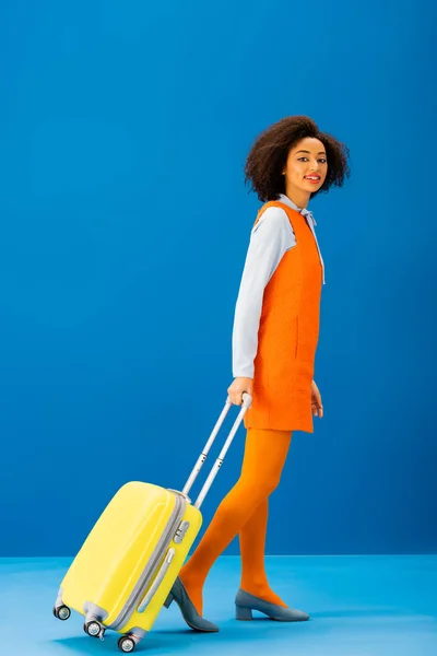 Улыбающийся африканский американец в ретро-платье ходить с дорожной сумкой на синем фоне — стоковое фото