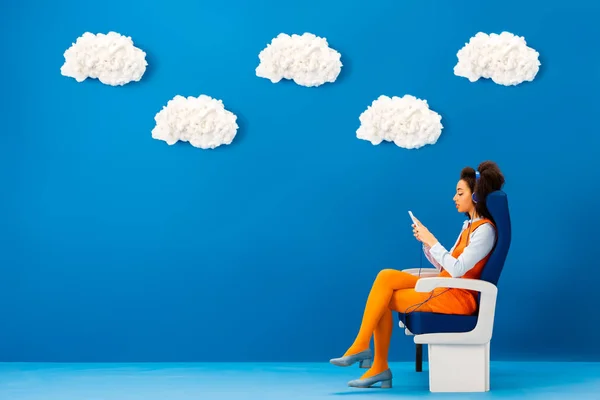 Seitenansicht eines Afroamerikaners im Retro-Kleid, der Musik hört und sein Smartphone auf blauem Hintergrund mit Wolken benutzt — Stockfoto