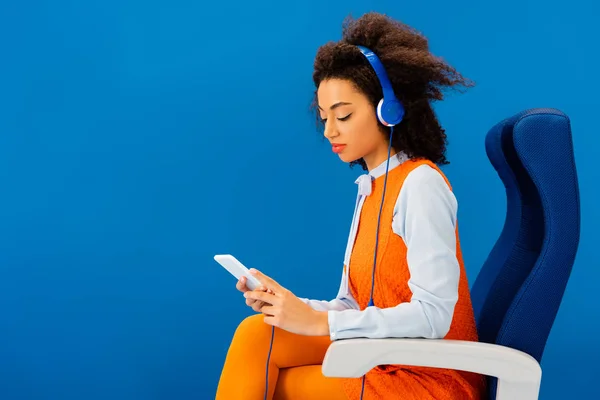 Afroamericano en vestido retro escuchando música y usando smartphone aislado en azul - foto de stock