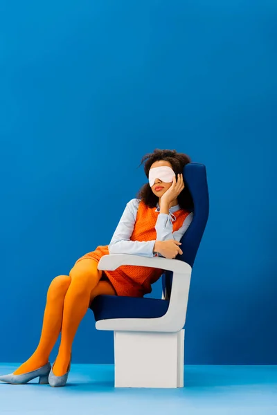Afroamericano con máscara de dormir sentado en el asiento y durmiendo sobre fondo azul - foto de stock
