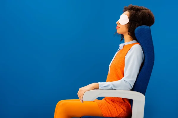 Side view of african american with sleeping mask sitting on seat and sleeping isolated on blue — Stock Photo