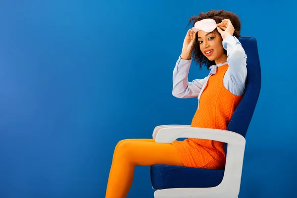 Souriant afro-américain en robe rétro tenant masque de sommeil et assis sur un siège isolé sur bleu — Photo de stock