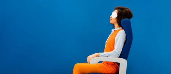 Panoramic shot of african american with sleeping mask sitting on seat and sleeping isolated on blue — Stock Photo