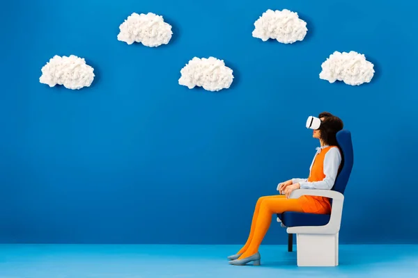 Vue latérale du casque afro-américain en vr assis sur un siège sur fond bleu avec des nuages — Photo de stock
