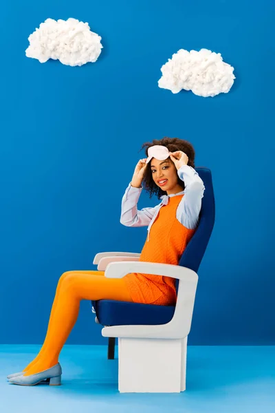 Smiling african american in retro dress holding sleeping mask and sitting on seat on blue background with clouds — Stock Photo